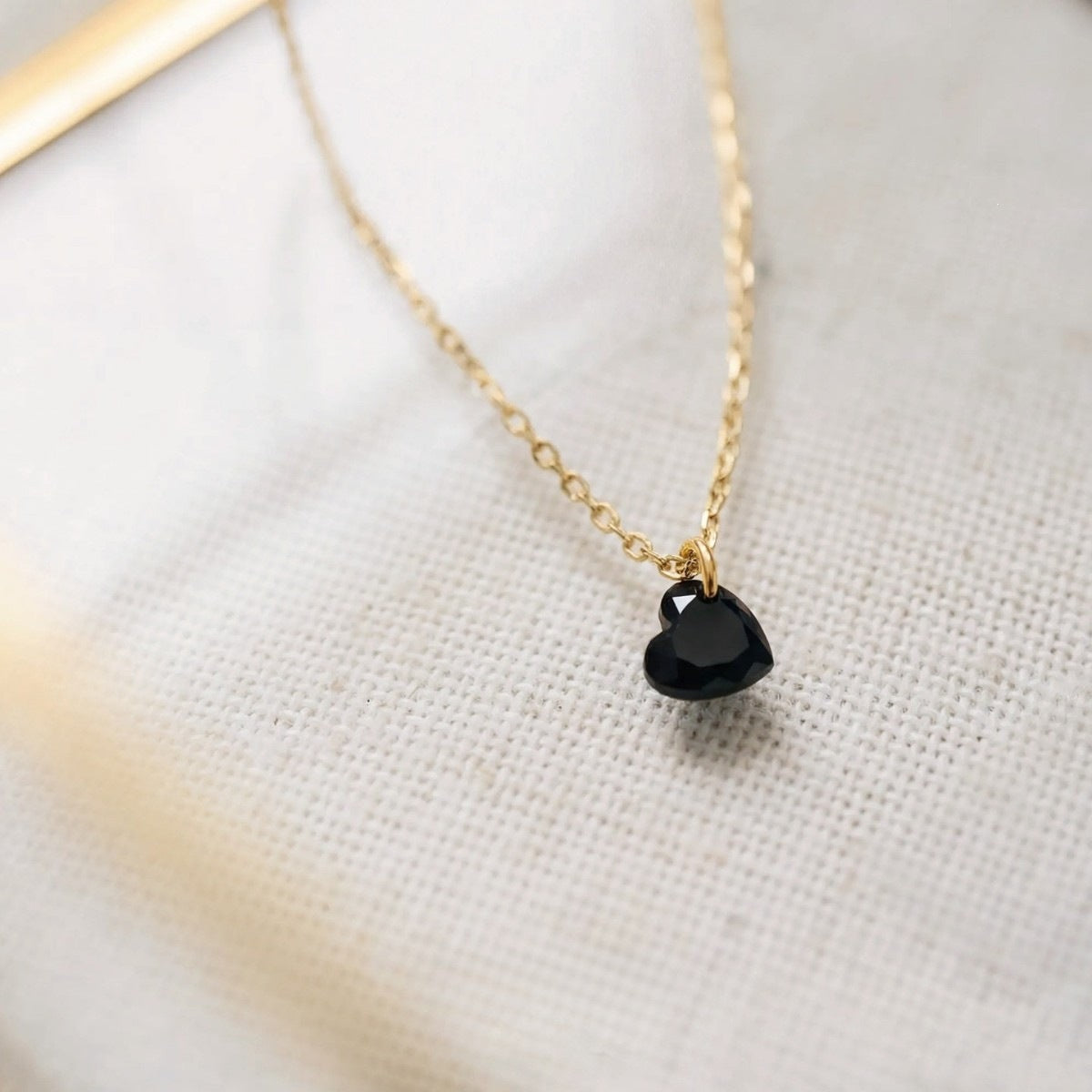 A delicate gold chain necklace featuring a small, polished black heart-shaped pendant, displayed on a textured off-white fabric background.