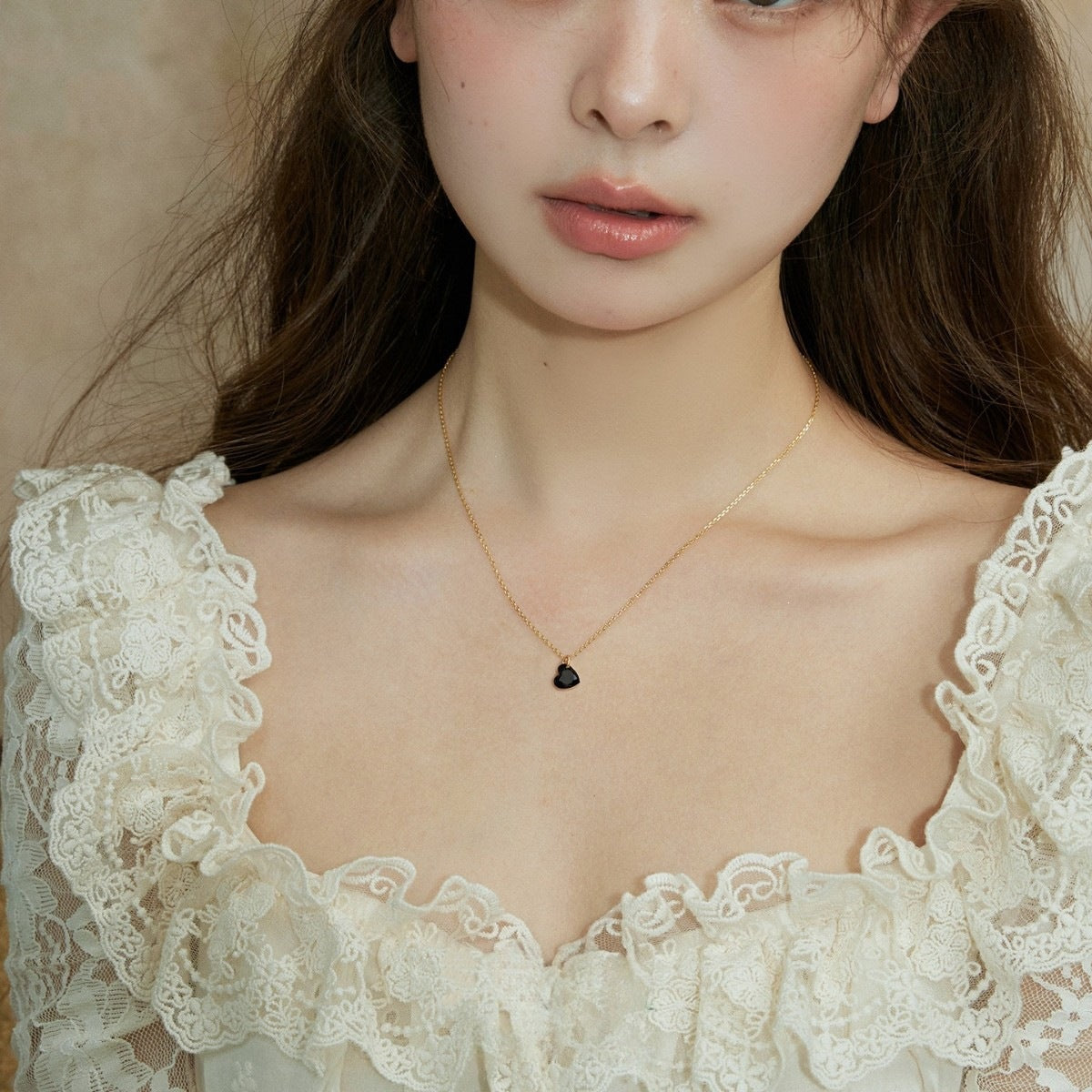 A close-up of a model wearing a delicate gold chain necklace with a small black heart-shaped pendant, paired with a cream lace top.