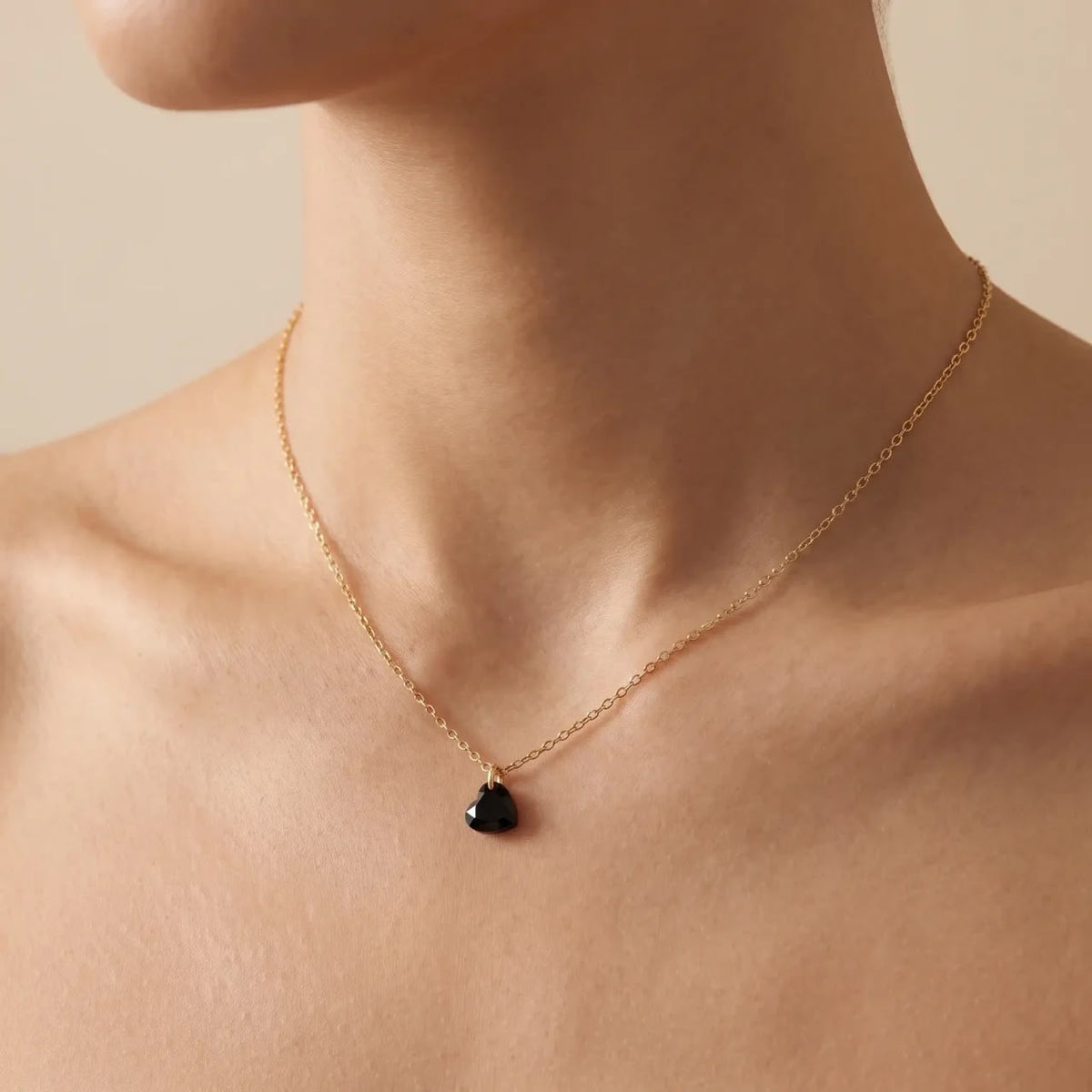 A delicate gold chain necklace with a small, polished black heart-shaped pendant, shown worn on a model's neck against a warm beige background.