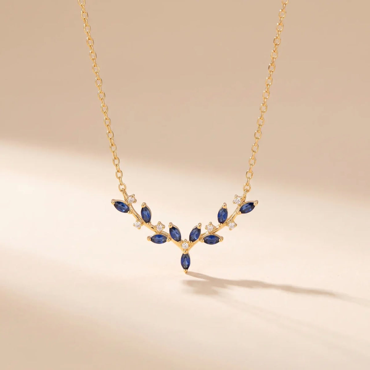 A gold V-shaped necklace with marquise-cut blue gemstones and small clear accent stones, displayed on a warm beige background.