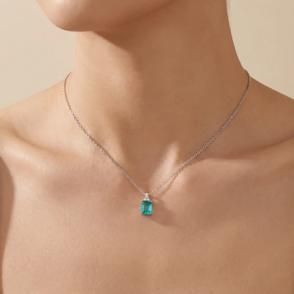 A close-up of a model's neck and upper chest wearing a delicate silver chain necklace with a rectangular emerald-cut turquoise pendant.