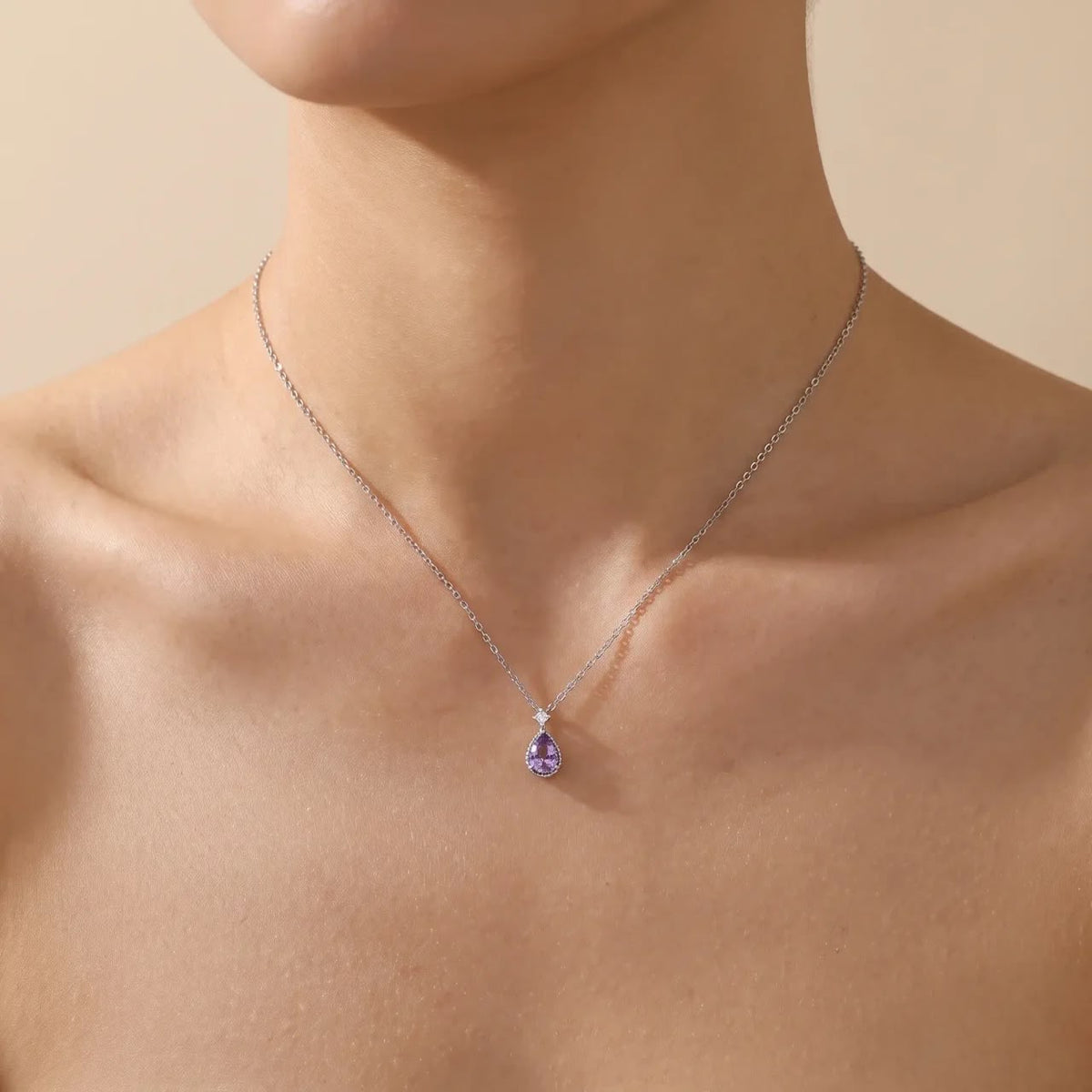 A close-up of a model's neck and upper chest wearing a delicate silver chain necklace with a small teardrop-shaped purple gemstone pendant.