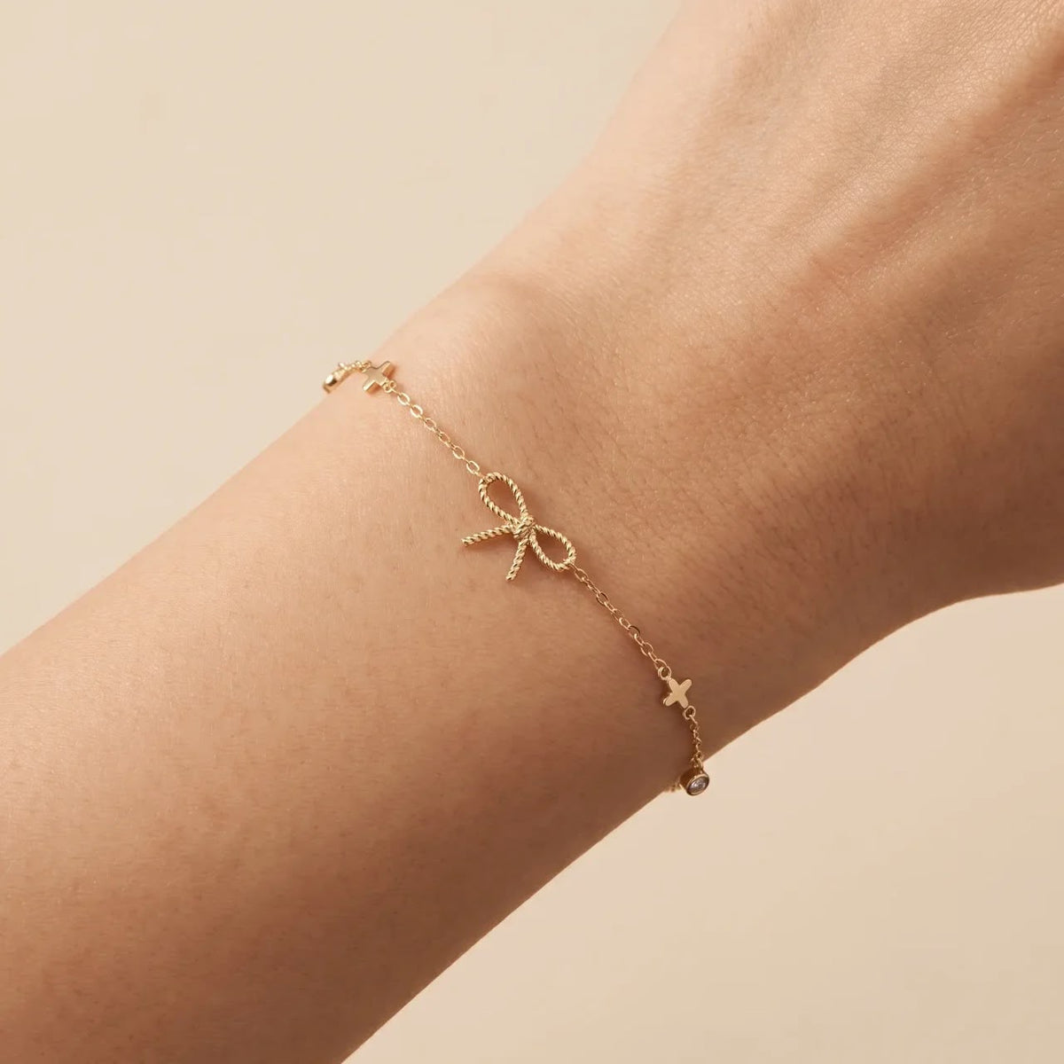 A delicate gold chain bracelet with a small bow charm and tiny star accents, shown worn on a person's wrist against a soft beige background.
