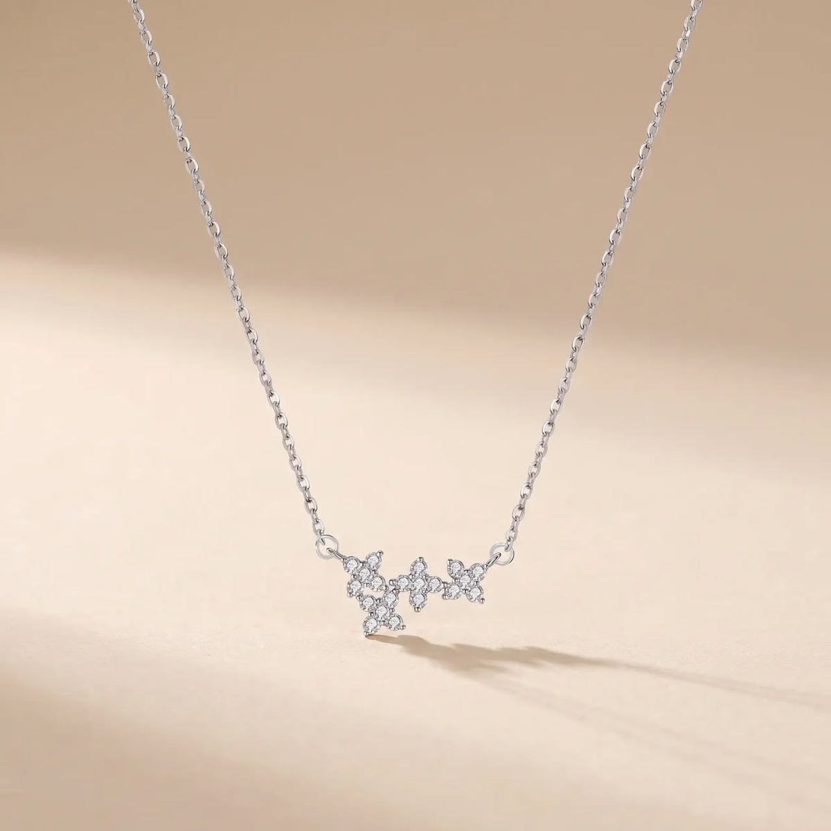 A delicate silver chain necklace featuring a small pendant composed of clear gemstones arranged in a butterfly-like silhouette, displayed against a soft beige background.