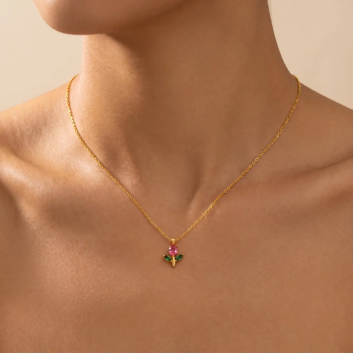 A close-up of a model's neck and upper chest wearing a delicate gold chain necklace with a small floral pendant featuring pink and green stones.