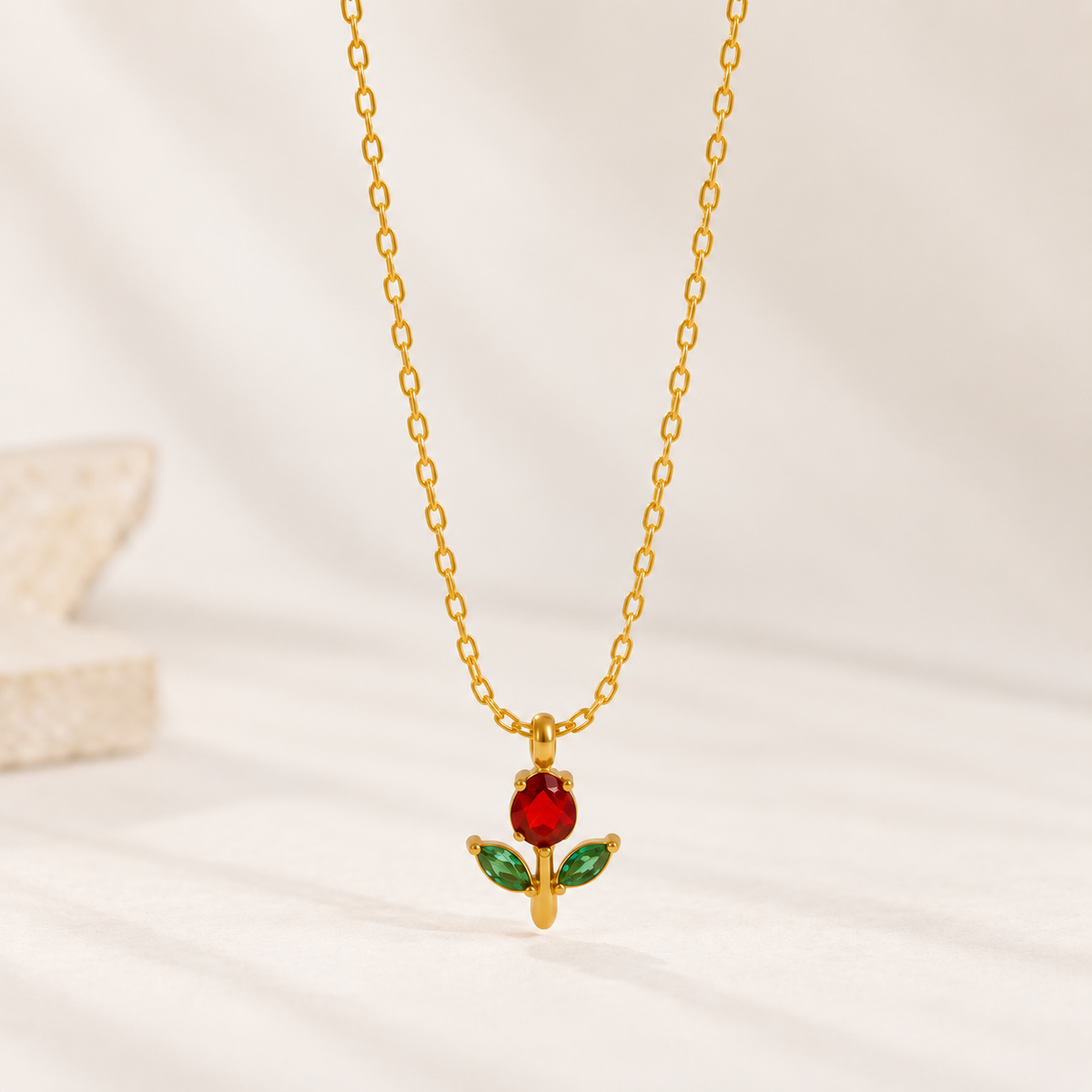A gold chain necklace with a pendant featuring a red rose bloom and green leaves, displayed on a soft white fabric background.