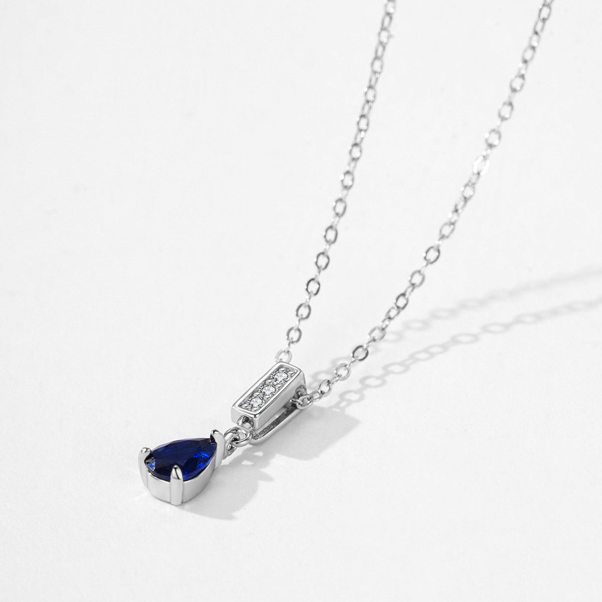 A silver chain necklace with a pear-cut blue gemstone pendant and a small rectangular pavé accent, photographed on a clean white background.