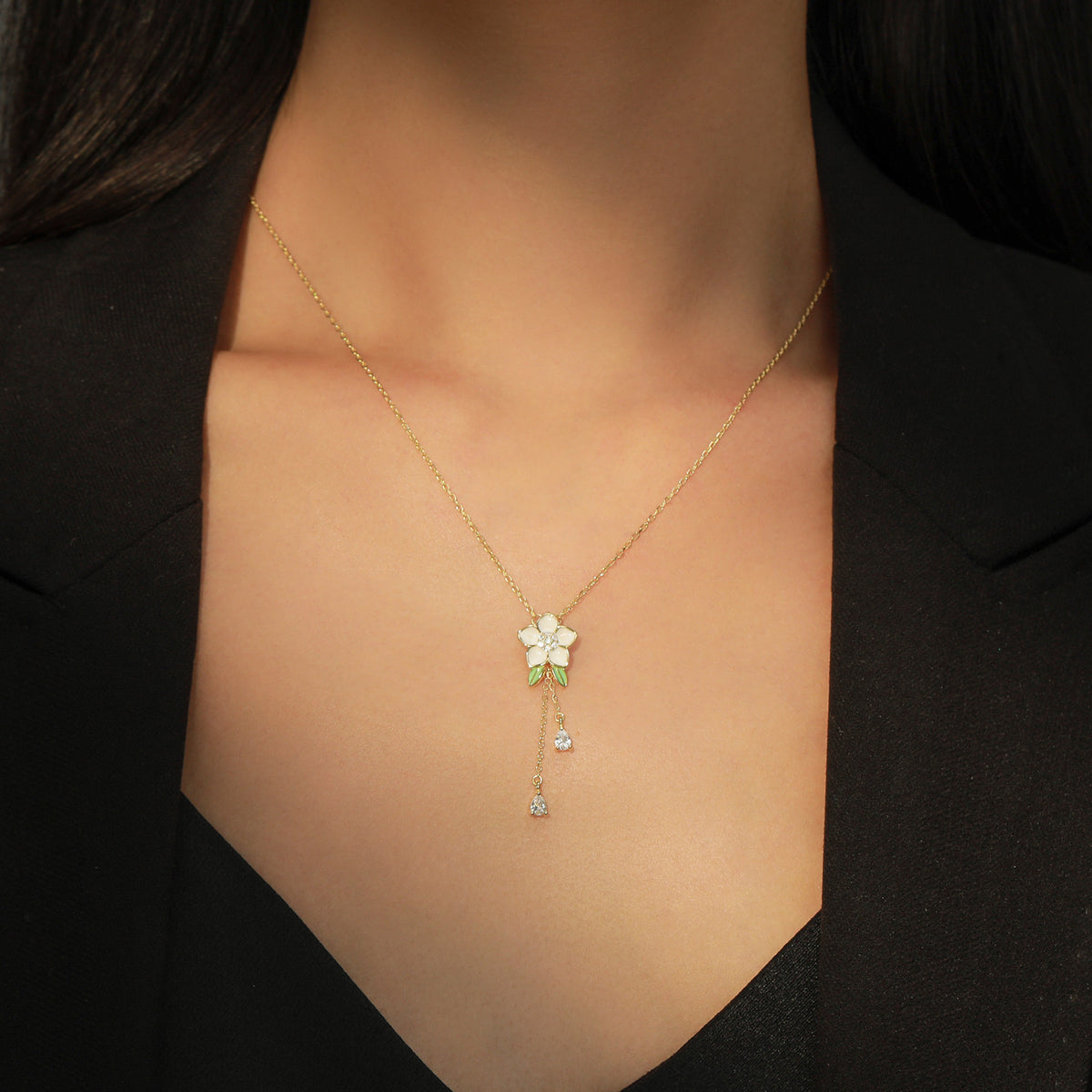 A delicate gold necklace with a white flower pendant and green leaves, worn by a model in a black blazer.